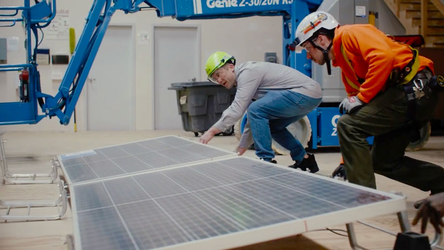 Two individuals wearing helmets are installing solar panels indoors with a blue lift in the background and industrial equipment around them.