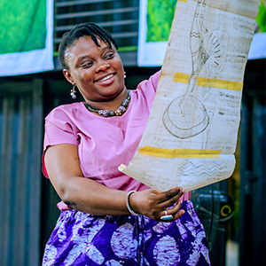 A smiling Nigerian woman in brightly colored clothing, holds a piece of artwork