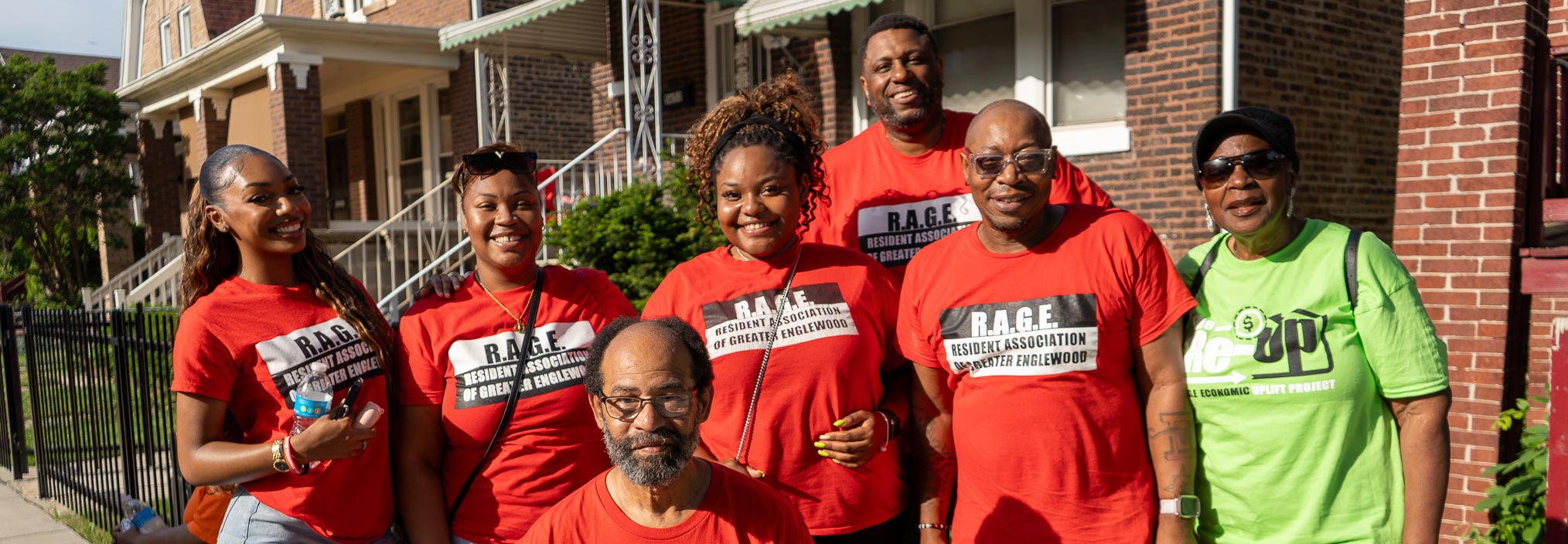 Seven people smiling outside, six in red â€œR.A.G.E.â€ shirts, one in a green shirt, standing in front of brick houses.